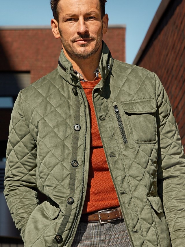 Ein Mann trägt eine 246529 Leichtsteppjacke in khaki, darunter einen Pullover in orange und dazu eine passende Hose in khaki.