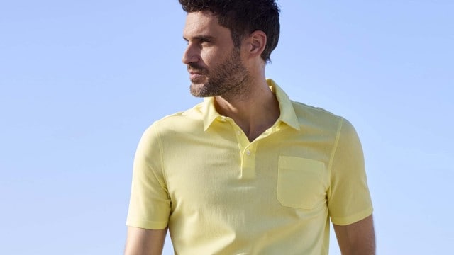 Ein Mann steht am Strand, trägt ein gelbes Poloshirt und blaue Jeans, und schaut zur Seite.