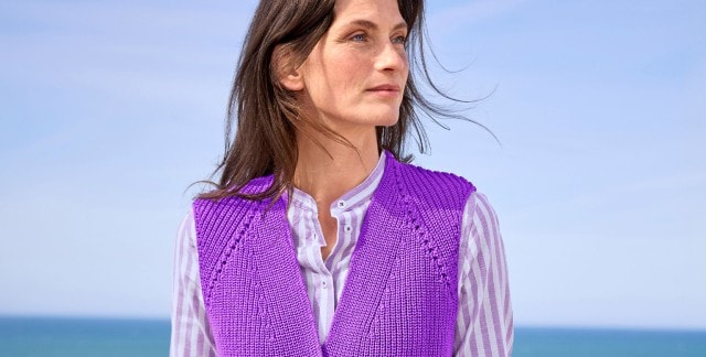 Eine Frau trägt eine gestrickte, lila Weste über einer lila-weiß gestreiften Bluse. Der Stil wirkt modern und farbenfroh.