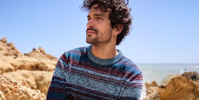 Ein Mann trägt einen gestreiften Strickpullover in Blau-, Rot- und Grautönen. Sein Stil wirkt modern und gleichzeitig entspannt.