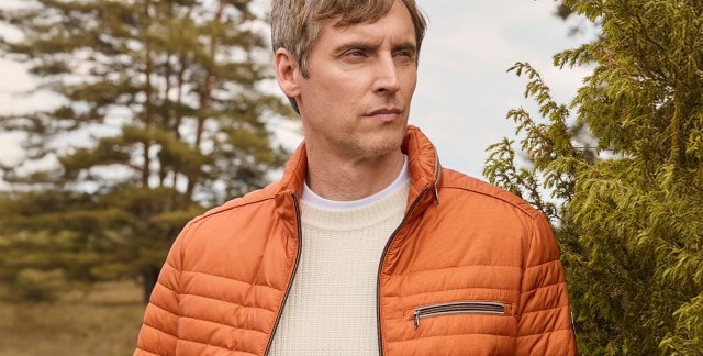 Ein Mann trägt eine Mircofaser Leichtjacke in Orange, darunter einen wollweißen Strickpullover.