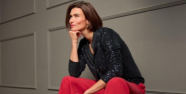 Eine Frau sitzt auf einem Stuhl, ist nach vorne gelehnt und trägt eine schwarze Paillettenjacke, dazu eine rote Hose.