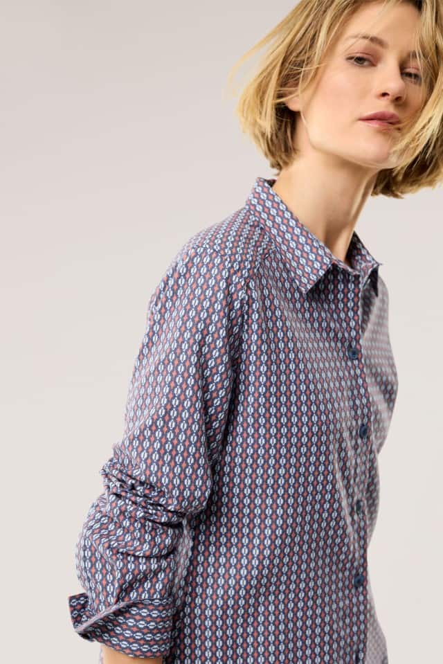 Eine Frau trägt ein langärmliges Hemd mit kleinem, geometrischem Muster in Blau-, Weiß- und Rottönen, das einen modernen, legeren Stil ausstrahlt.