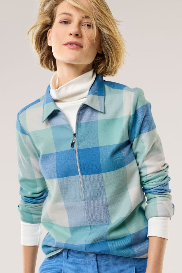Eine Person trägt ein Oberteil mit großem Karomuster in Blau-, Grün- und Weißtönen, mit Reißverschluss am Kragen und weißem Rollkragen darunter, kombiniert mit einer hellblauen Hose für einen lässigen, modernen Look.