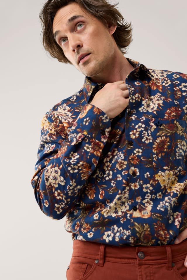 Eine Person trägt ein blaues Hemd mit floralem Muster in warmen Farbtönen und eine rostrote Hose im modisch-lässigen Stil.