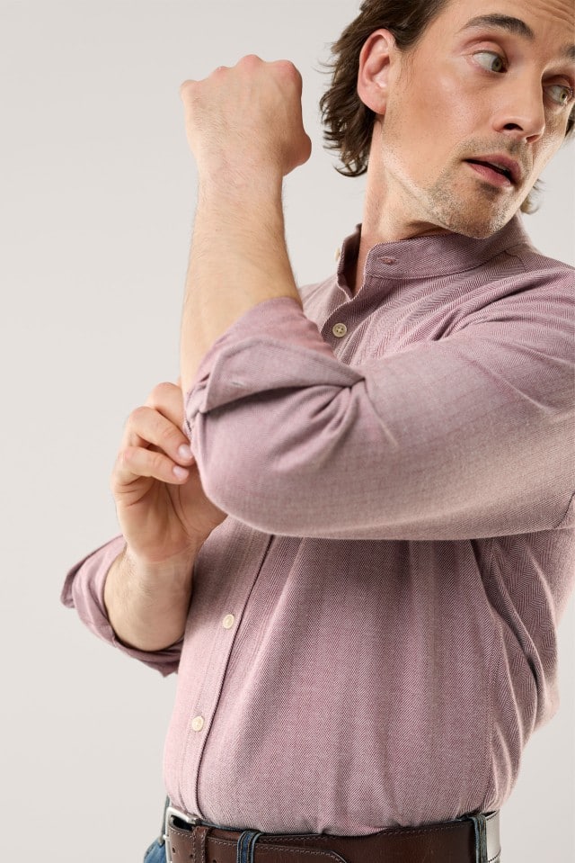 Eine Person trägt ein hellrosafarbenes Hemd mit dezentem Fischgrätmuster und krempelt lässig die Ärmel hoch, kombiniert mit Jeans und braunem Gürtel.