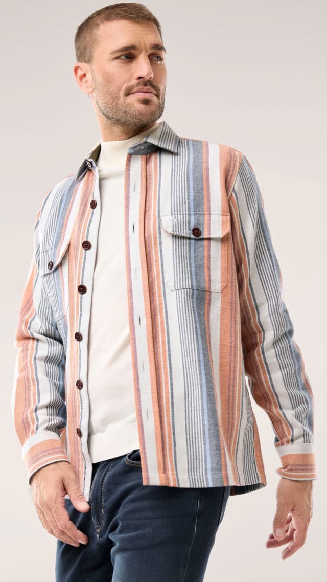 Eine Person trägt ein langärmliges Hemd mit breiten Streifen in Orange-, Blau- und Beigetönen über einem hellen Rollkragenpullover, kombiniert mit einer dunklen Jeans für einen entspannten Look.