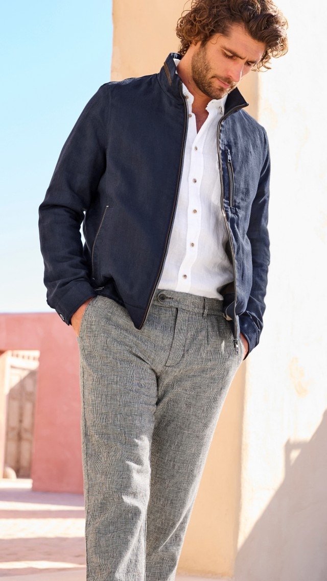 Ein Mann trägt eine dunkelblaue, leichte Jacke über einem weißen Leinenhemd und kombiniert dazu eine graue Hose mit feinem Karomuster. Der Look ist modern, stilvoll und eignet sich gut für milde Tage.