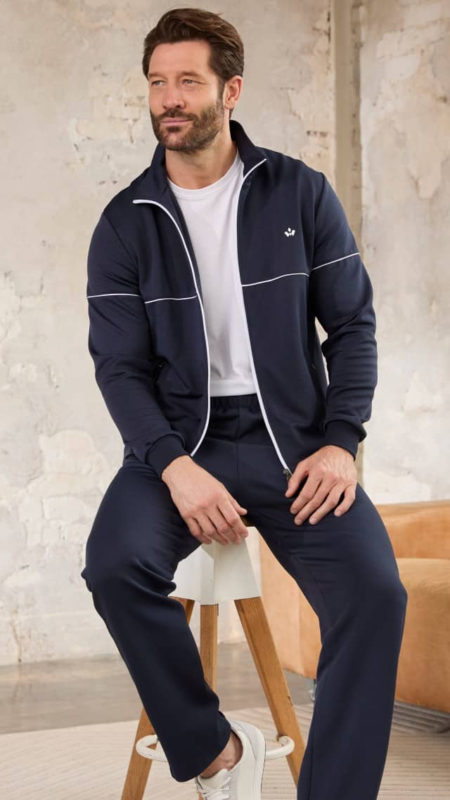 Ein Mann sitzt entspannt auf einem Hocker und trägt einen dunkelblauen Freizeitanzug mit weißen Akzenten sowie ein schlichtes weißes T‑Shirt. Der Look wirkt sportlich, modern und bequem.