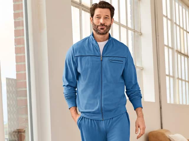 Ein Mann trägt einen blauen Freizeitanzug mit Zip‑Jacke und passender Hose in einem hellen, modernen Innenraum. Der Look wirkt sportlich, lässig und bequem.