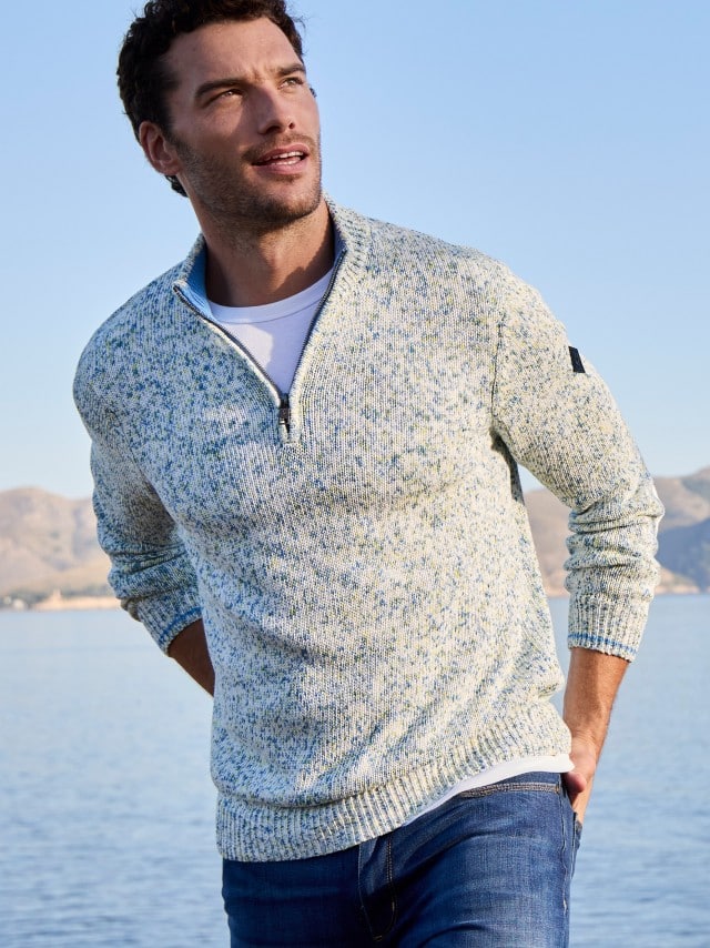Ein Mann steht am Wasser und trägt einen hell melierten Troyer‑Pullover über einem weißen T‑Shirt sowie eine Jeans.