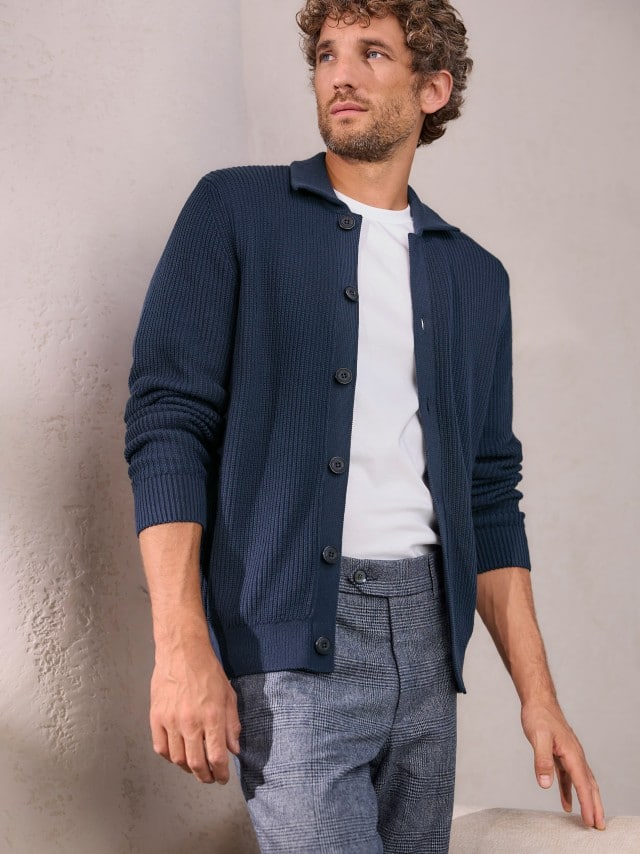 Ein Mann trägt einen gerippten Polo-Cardigan in Marine, darunter ein weißes Shirt und dazu eine passende Hose in Blau.