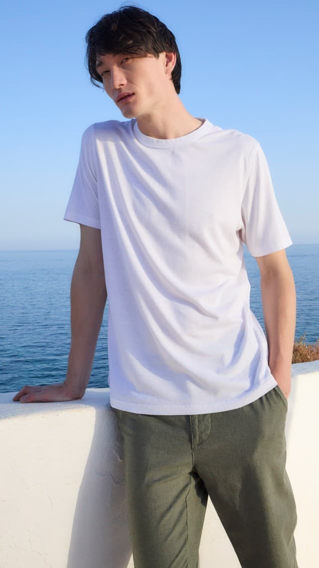 Ein Mann lehnt an einer weißen Mauer am Meer und trägt ein weißes T‑Shirt sowie eine olivgrüne Hose.