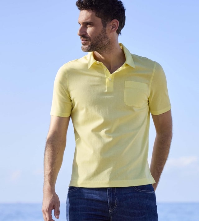 Ein Mann steht am Strand, trägt ein gelbes Poloshirt und blaue Jeans, und schaut zur Seite.