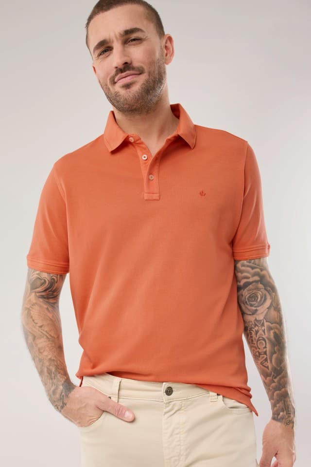 Ein Mann trägt ein Polo-Shirt in Orange und hält seine Hand in  der Hosentasche.