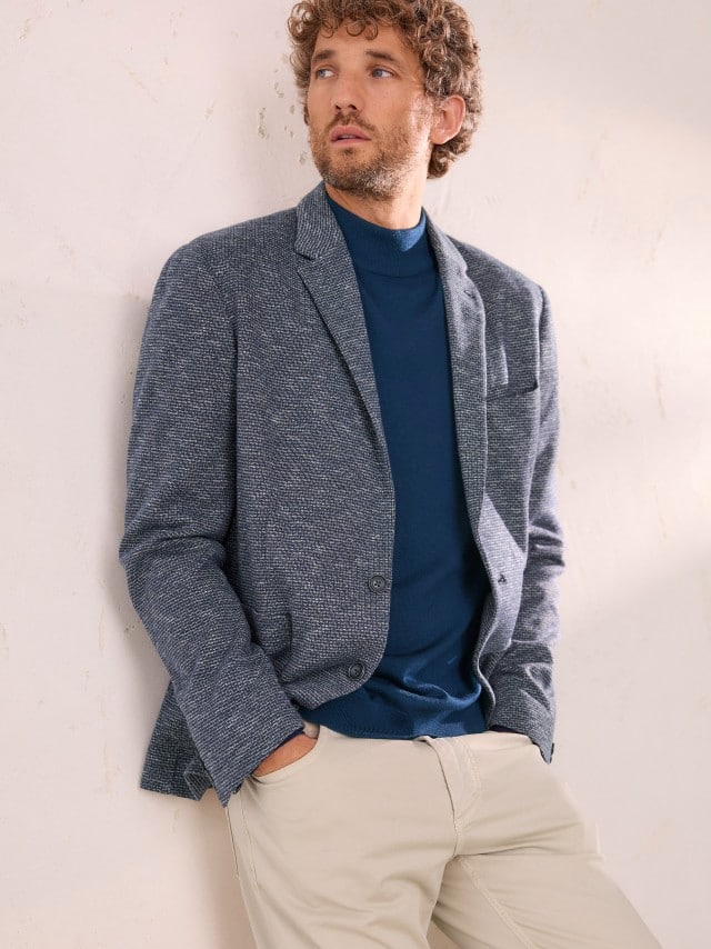 Ein Mann an einer Wand gelehnt, trägt ein blaues High-Flex Sakko, darunter einen Pullover in Marine und dazu eine Hose in Beige. 