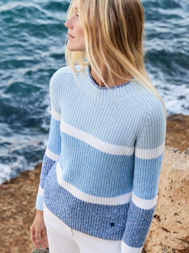 Eine Frau steht am Meer und trägt einen hellblau‑weiß gestreiften Strickpullover zu einer weißen Hose.