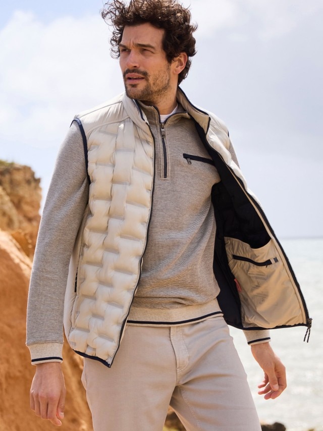Ein Mann trägt einen hellgrauen Pullover mit Zipper und darüber eine gesteppte, ärmellose Weste. Dazu kombiniert er eine beigefarbene Hose für einen sportlich-eleganten Look.