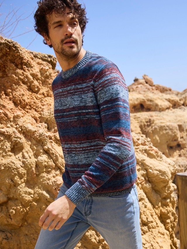 Ein Mann trägt einen gestreiften Strickpullover in Blau-, Rot- und Grautönen. Dazu kombiniert er eine hellblaue Jeans für einen legeren, modernen Look.