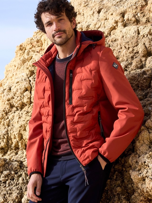 Ein Mann trägt eine rote Steppjacke mit Kapuze über einem passenden Pullover. Dazu kombiniert er eine dunkelblaue Hose für einen sportlich-lässigen Look.