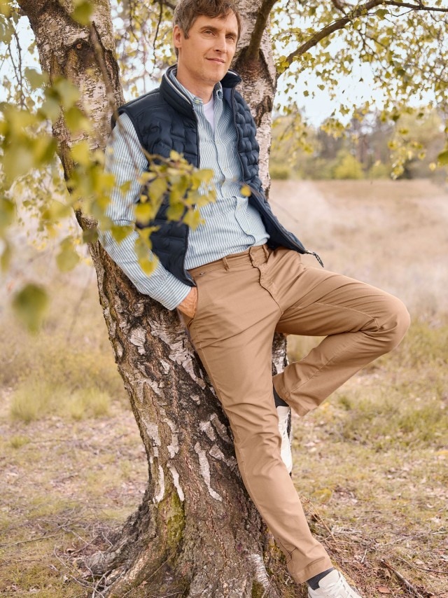 Ein Mann am Baum angelehnt trägt eine Leichtweste in Marine, darunter ein gestreiftes Hemd und dazu eine Hose in Beige.