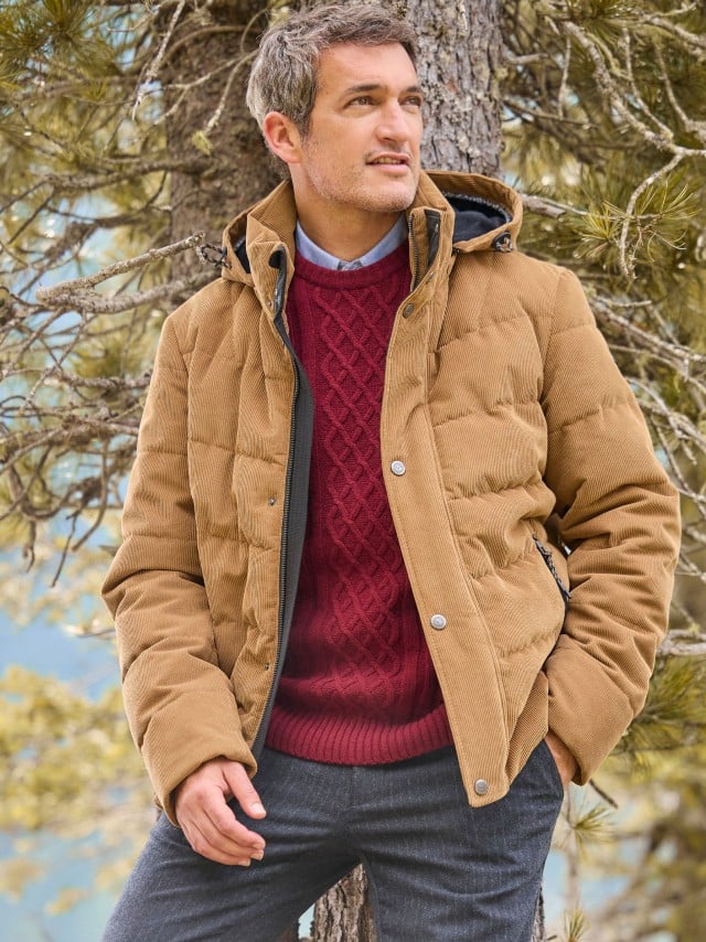 Ein Mann steht vor einem Baum und trägt die Cord Steppjacke in Camel, darunter einen roten Strickpullover mit Hemd und dazu eine Sport-Flanell Hose.