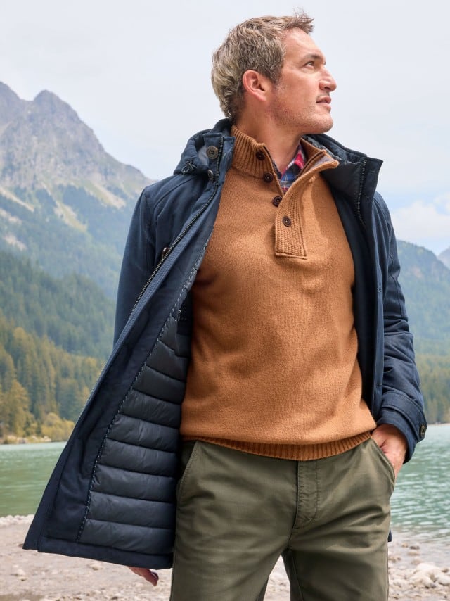 Ein Mann steht vor einem Gebirge, schaut zur Seite und trägt den Ultraskin Leichtparka in Marine, darunter einen Premium Lammwoll-Troyer in Camel und dazu eine Hose in Oliv.