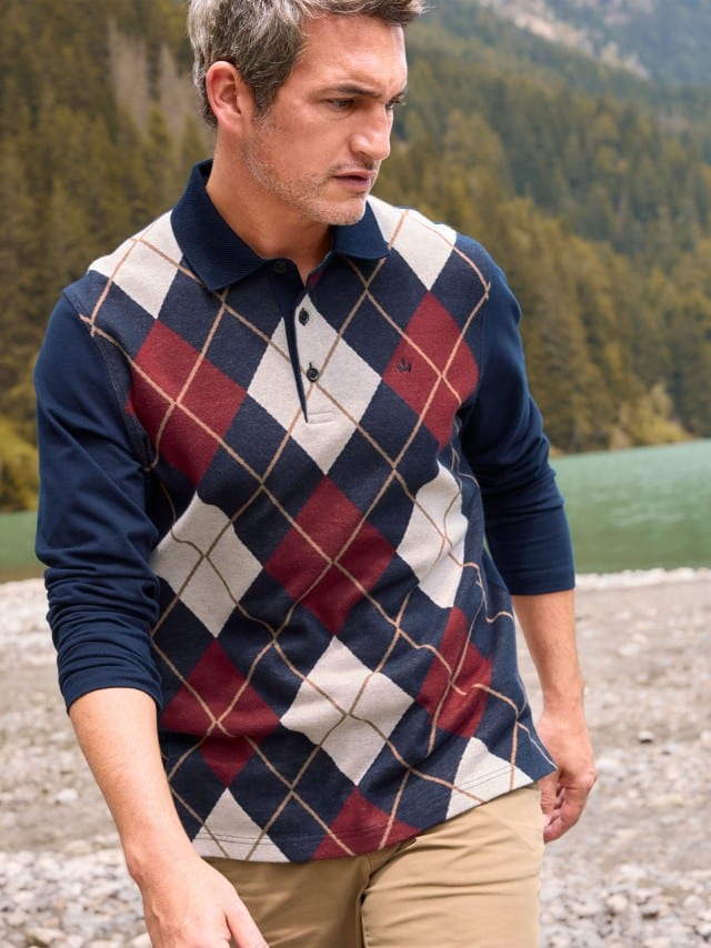 Ein Mann steht vor einem Bergsee und trägt ein Rauten-Polo Supersoft, dazu eine Hose in Beige. 