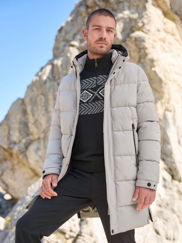 Ein Mann steht vor einem Berg und trägt den Thermo Steppmantel in Ecru, dazu einen Norweger Troyer in Schwarz und eine schwarze Hose.