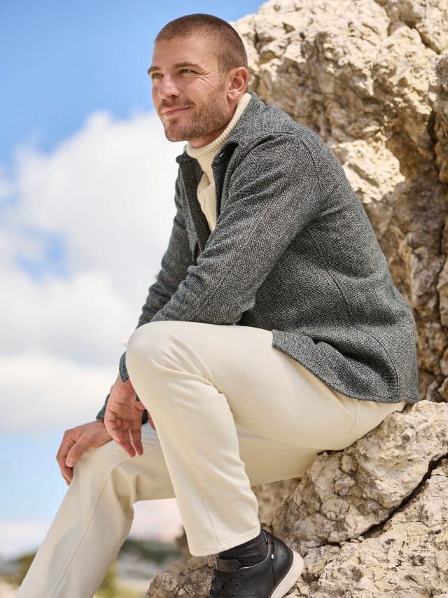 Ein Mann hockt auf Felsen und trägt eine Hemd-Jacke in Schwarz/Weiß, dazu einen Rollkragen-Pullover mit Zopfmuster in Wollweiß und eine weiße Hose.