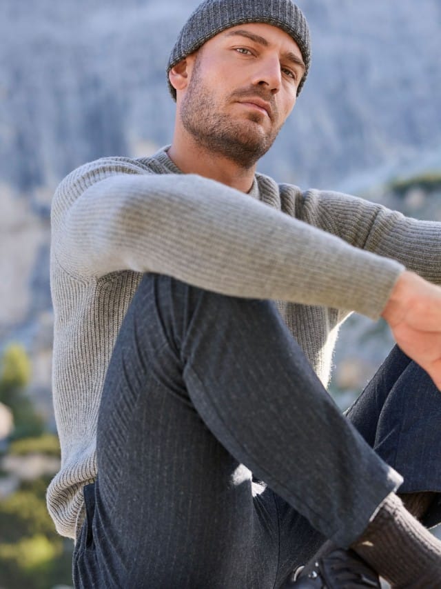 Ein Mann trägt einen grauen gerippten Strickpullover, dazu eine Premium Sport-Flanell Hose in Anthrazit gestreift und eine passende Mütze.