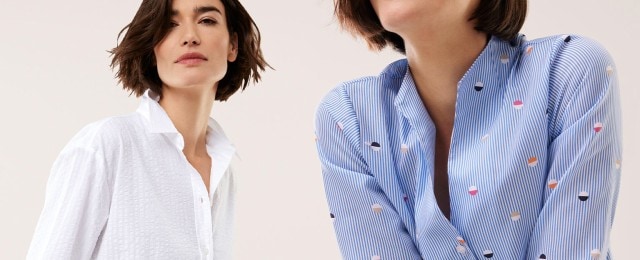 Zwei Frauen präsentieren Blusen: eine trägt eine schlichte weiße Bluse, die andere eine blau-weiß gestreifte Bluse mit buntem Muster. Beide Outfits wirken modern und frisch.