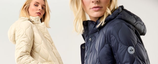 Zwei Frauen tragen gesteppte Kapuzenjacken: Eine in hellem Beige, die andere in Dunkelblau mit Logo-Detail am Ärmel. Die Jacken wirken modern, sportlich und sind ideal für kühle Tage geeignet