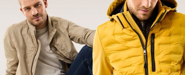 Ein Mann trägt eine lässige, beige Jacke, der andere eine auffällige, gelbe Steppjacke. Beide Looks strahlen modernen, individuellen Stil aus.