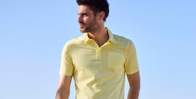 Ein Mann steht vor blauem Himmel und trägt ein kurzärmliges, hellgelbes Poloshirt mit Kragen und Brusttasche.