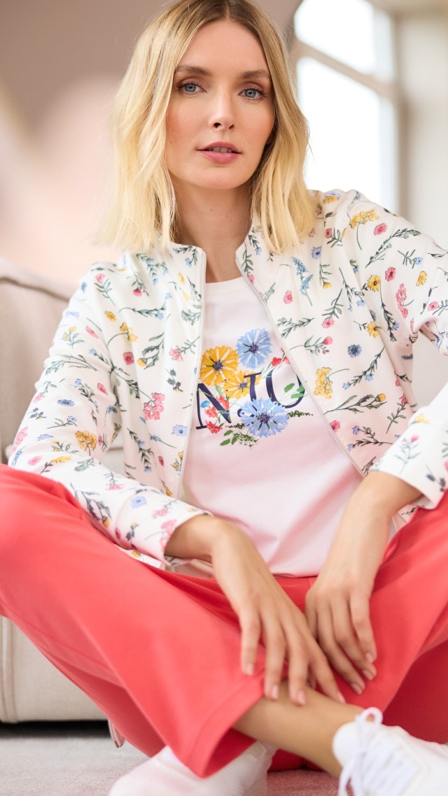 Blonde Frau im Schneidersitz in geblümter Freizeitjacke, Shirt und koralle farbener Hose.