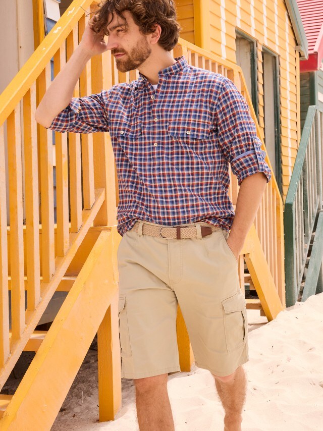 Ein Mann mit braunen Haaren steht barfuß im Sand vor einer gelben Holztreppe, trägt ein kariertes Hemd und beige Shorts und schaut nachdenklich zur Seite.