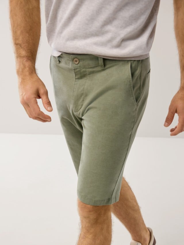 Ein Mann trägt eine hellgraue T-Shirt und eine grüne kurze Hose; das Bild zeigt ihn von der Hüfte abwärts beim Gehen in einem hellen Raum.