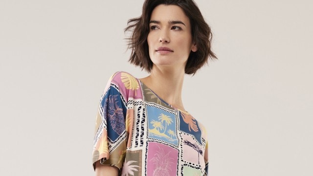 Eine Frau steht in einem hellen Raum, trägt ein bunt gemustertes Shirt und einen weiten hellblauen Rock. 