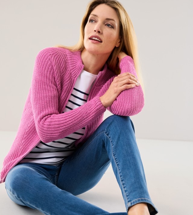 Frau in einem gestreiften Oberteil und rosa Strickjacke sitzt vor einem neutralen Hintergrund.