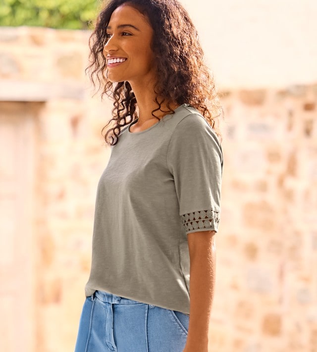 Frau trägt ein grünes T-Shirt und Jeans und steht seitlich vor einer Steinmauer.