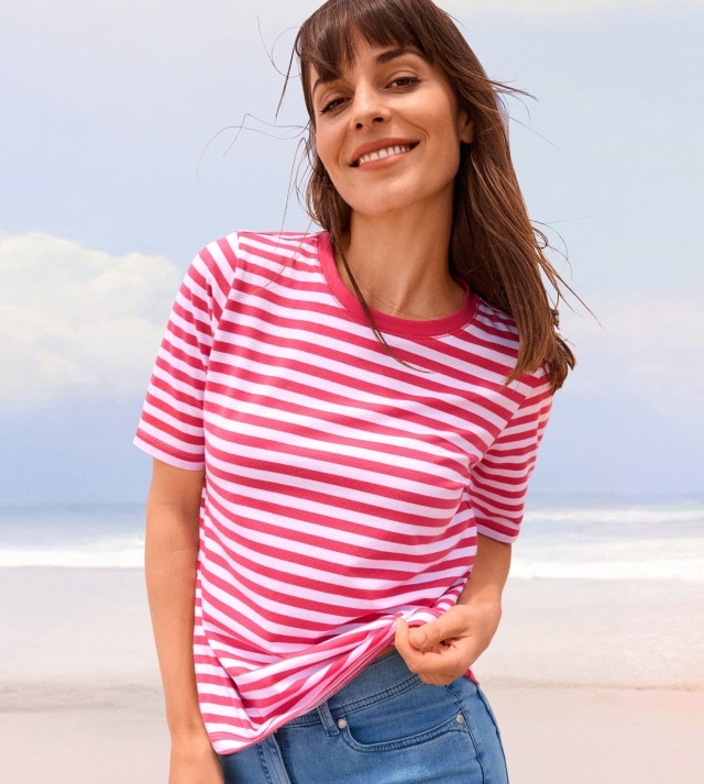 Frau in einem rot-weiß gestreiften T-Shirt und Jeans steht am Strand.