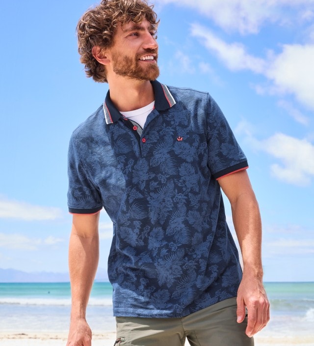 Ein Mann trägt ein dunkelblaues Poloshirt mit Muster und eine olivgrüne Hose und steht am Strand.