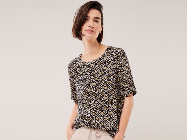 Frau trägt ein beige-braun gemustertes Shirt und steht vor einer hellen Wand