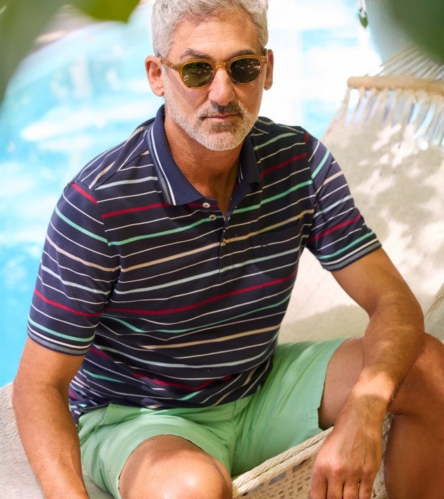 Ein Mann sitzt in einer Hängematte am Pool und trägt ein dunkelblaues Poloshirt mit bunten, schmalen Querstreifen sowie eine hellgrüne Shorts.