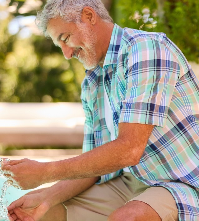 Ein Mann sitzt am Pool, spielt mit Wasser und trägt ein kurzärmliges, kariertes Hemd in verschiedenen Blau-, Grün- und Weißtönen sowie eine beige Shorts.
