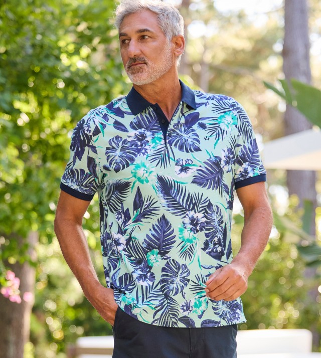 Ein Mann steht im Grünen und trägt ein kurzärmliges Poloshirt mit tropisch-blauem Blumen- und Blätterdruck auf hellem Hintergrund.