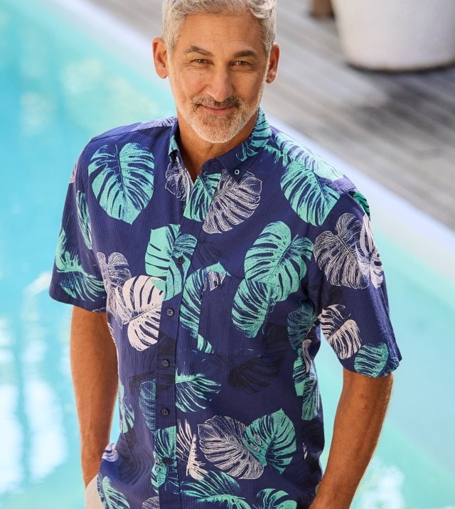 Ein Mann sitzt am Pool und trägt ein blaues Leinenhemd mit Blattmuster. 