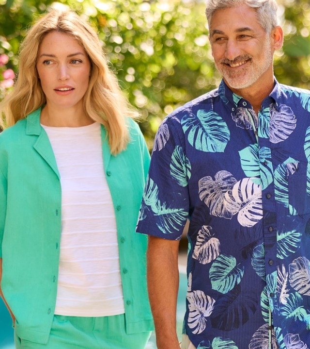 Ein Mann und eine Frau stehen nebeneinander im Freien; die Frau trägt eine helle, türkisfarbene Sommerjacke mit passender Hose und weißem T-Shirt, der Mann ein blaues, kurzärmliges Hemd mit hellem Blätterdruck.
