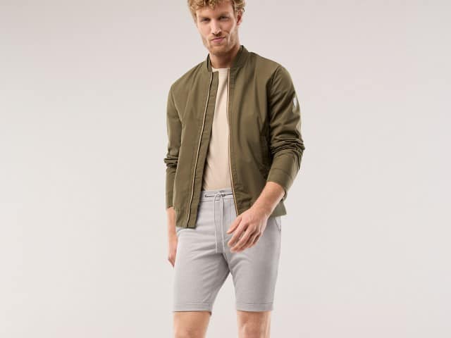Ein Mann trägt eine Jersey Shorts in der Farbe Sand, dazu eine helles Shirt mit Blouson.